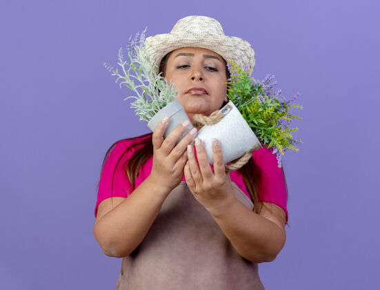 帽子穿着围裙 戴着帽子 手持盆栽植物的中年园丁站在紫色背景下 带着怀疑的表情看着它们抱着站着表情