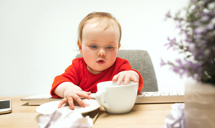 小快乐的小女婴坐在杯子和键盘的现代电脑或笔记本电脑隔离在白色笔记本电脑桌子幼儿