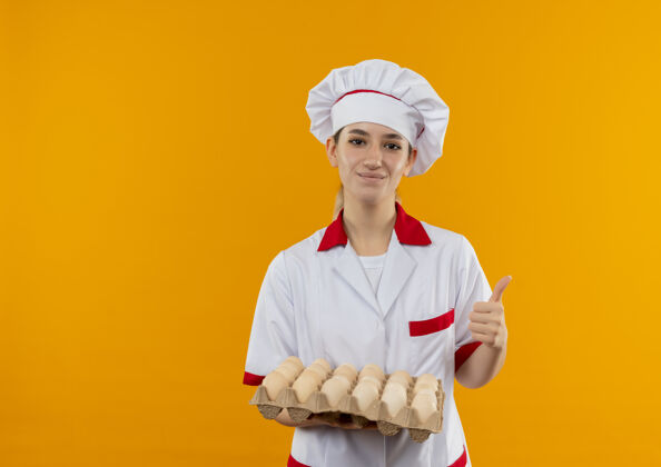 橙色年轻漂亮的厨师穿着厨师制服 手里拿着一盒鸡蛋 在橙色的空间里孤立地竖起大拇指年轻鸡蛋拇指