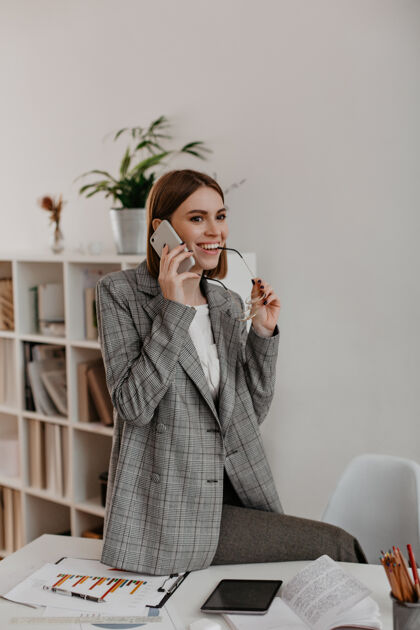 开朗开朗的商务女士微笑着通过电话交流穿着灰色格子夹克的女士在白色办公室摆姿势成功专业人士灰色夹克