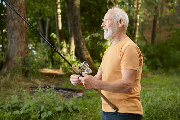 老年人帅气的欧洲男性退休老人在松树上钓鱼的侧视图 拿着钓竿从水里捞上来 开心地微笑着 在野外享受着活跃的户外爱好捕获垂钓者户外