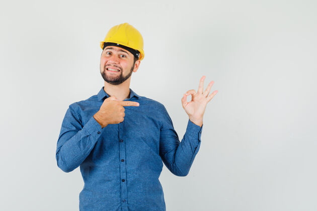 仓库一个穿着衬衫 戴着头盔的年轻工人指着他的ok标志 看上去很高兴沟通头脑风暴工作