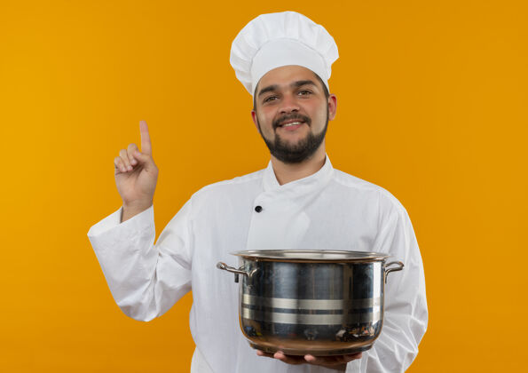 微笑面带微笑的年轻男厨师身着厨师制服 手拿锅子 抬起头孤立地看着橙色的空间向上拿着看