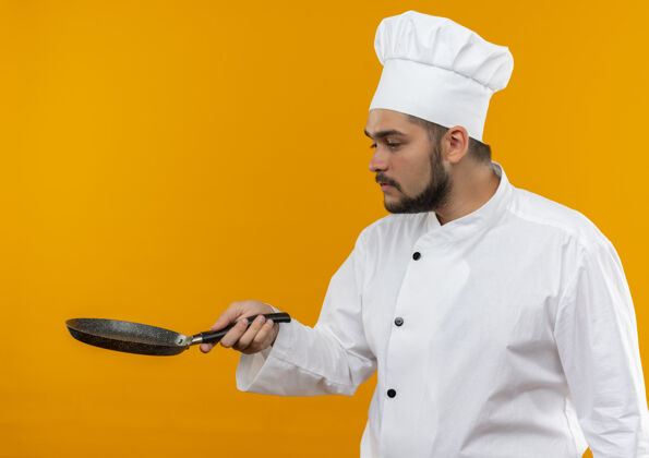 薯条年轻的男厨师身着厨师制服 手拿煎锅 看着孤立的橙色空间烹饪制服厨师