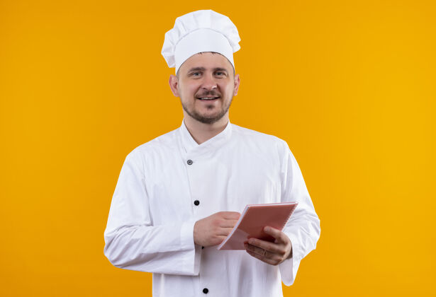 微笑微笑着的年轻帅哥厨师穿着厨师制服拿着笔记本孤立在橙色的空间橙色厨师垫着