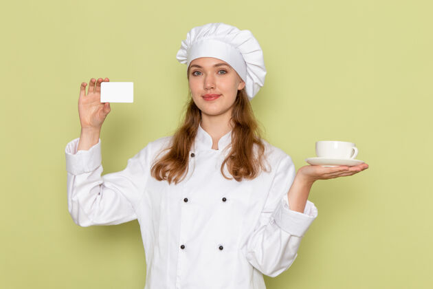 人身着白色厨师服的女厨师手持绿色墙壁上的杯子和卡片的正视图护士杯子女厨师