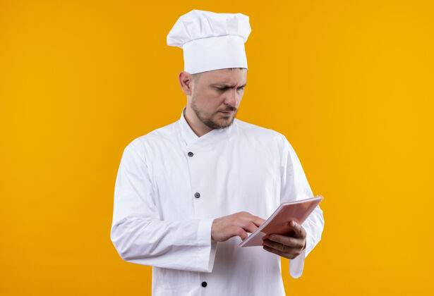 垫着体贴的年轻帅哥厨师穿着厨师制服拿着便笺簿把手放在上面 看着它孤立在橙色的空间里烹饪笔记厨师