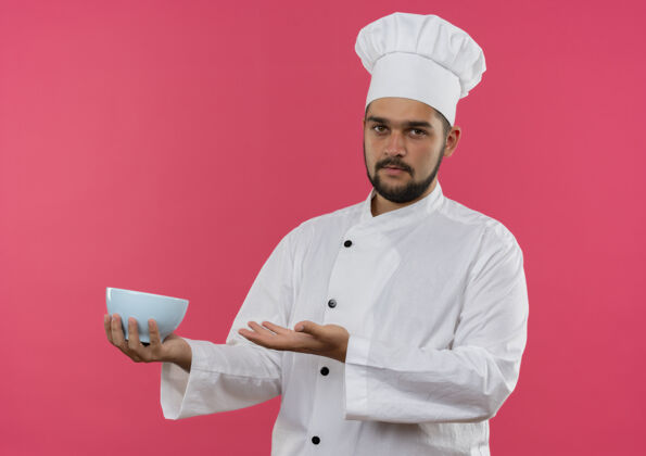 制服年轻的男厨师穿着厨师制服 拿着一个孤立的碗在粉红色的空间里年轻烹饪持有