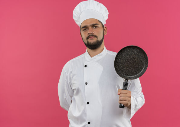 厨师穿着厨师制服的年轻男厨师手捧煎锅 一只手放在背后 被隔离在粉红色的空间里男性空间持有