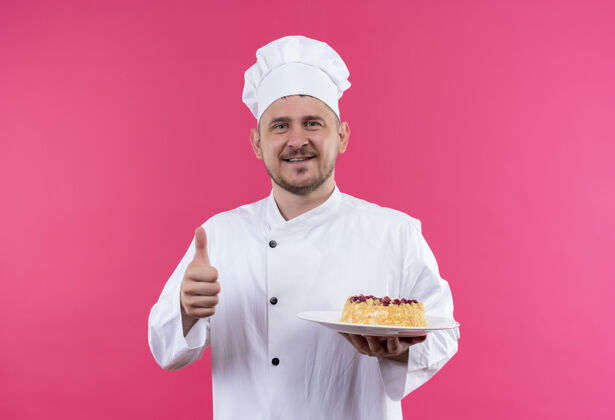 年轻面带微笑的年轻帅哥厨师身着厨师制服拿着一盘蛋糕在粉红色的空间里孤立地竖起大拇指拿着厨师蛋糕