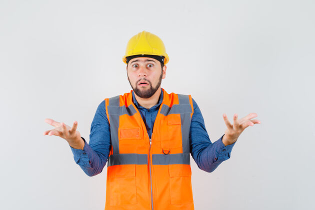 建筑年轻的建筑工人在衬衫 背心 头盔上表现出无助的姿态 看上去很困惑前视图肖像工作勤杂工