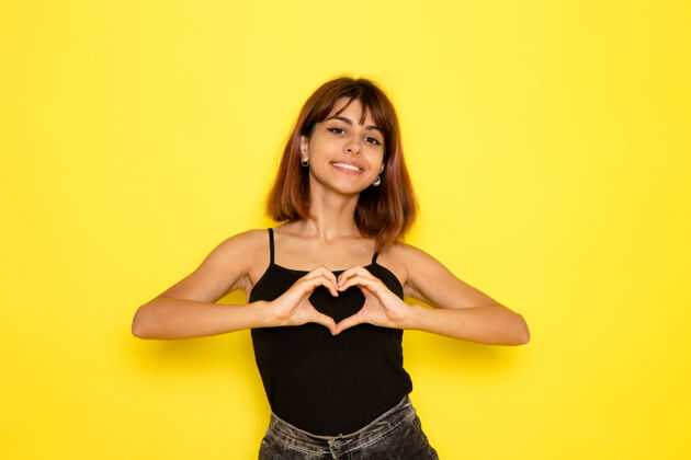 光身穿黑色衬衫的年轻女性在浅黄色墙上微笑并显示心形标志的正面视图姿势模型显示