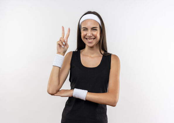 运动微笑的年轻漂亮的运动女孩戴着头带和腕带眨眼 并在白色空间做和平姿态年轻头带腕带