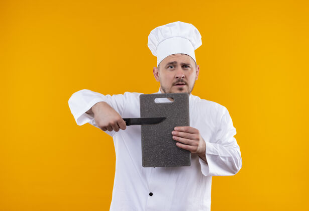 刀年轻帅气的厨师穿着厨师制服拿着砧板和刀隔离在橙色的空间里手持橘子切割