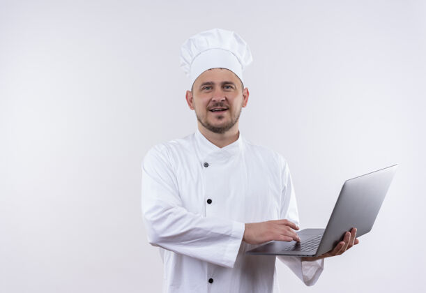 帅气高兴的年轻帅哥厨师穿着厨师制服拿着笔记本电脑在孤立的空白处年轻厨师持有