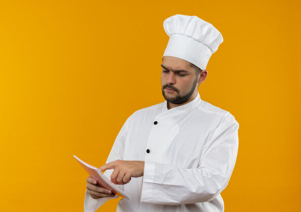 空间体贴的年轻男厨师穿着厨师制服 拿着手指看着并把手指放在隔离在橙色空间的记事本上厨师手指复制