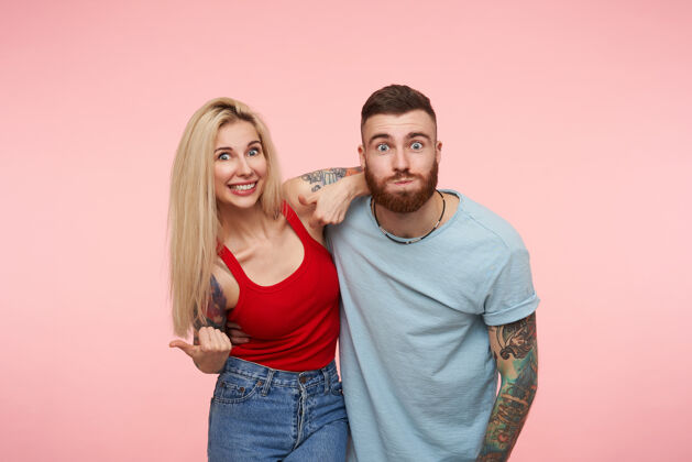 女士一对有纹身的积极的年轻漂亮的夫妇互相拥抱 一边做着滑稽的脸 一边兴奋地看着 站在粉红色的地板上休闲短裤双人