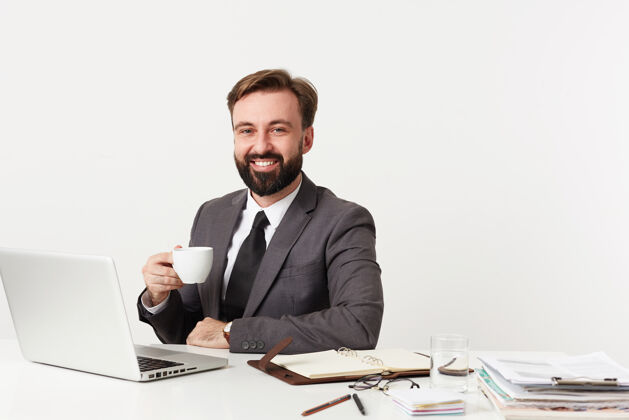 胡须照片中的是一位快乐英俊的年轻黑发男性 留着胡子 穿着灰色西装 打着白色的领带 一边喝着咖啡一边开心地微笑着肖像发型积极