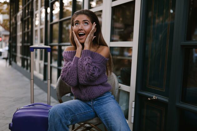 裙子退出模特带着蓝色手提箱坐在咖啡馆附近的椅子上 穿着紫色套头衫 牛仔裤 化妆 发型 情感 惊喜 秋天 金发 快乐 针织 微笑时尚手提箱迷人