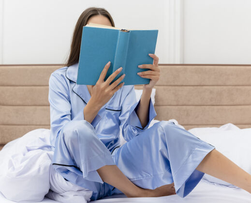 卧室穿着蓝色睡衣的年轻美女坐在床上 在卧室的室内灯光背景下看书床书室内