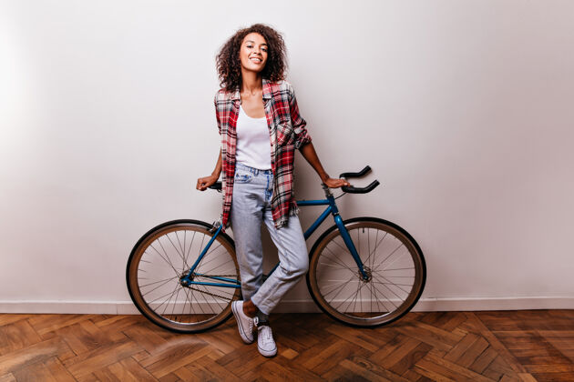 寒冷吸引人的女自行车手的全长肖像摄影棚拍摄的感兴趣的穿着牛仔裤的非洲女孩站在白色的自行车上时尚黑色衬衫