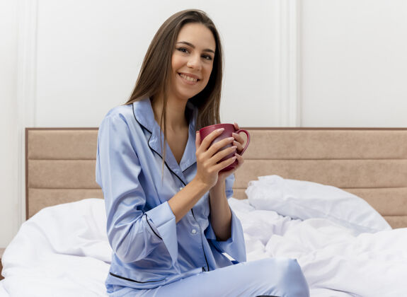 周末穿着蓝色睡衣的年轻漂亮女人坐在床上 喝着咖啡 看着相机 愉快而积极地微笑着 在明亮的背景下 在卧室里享受周末年轻休息杯子