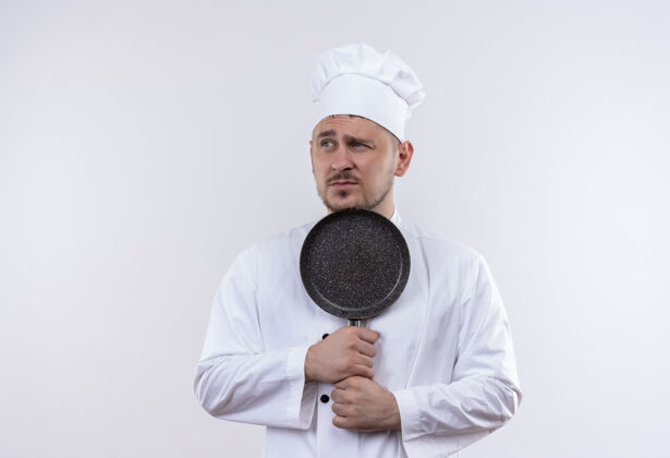 看年轻帅气的厨师穿着厨师制服 拿着煎锅 看着隔离在白色空间的一边不愉快厨师薯条