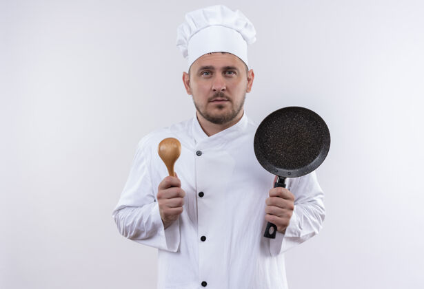 帅气严肃的年轻帅哥厨师穿着厨师制服拿着勺子和煎锅看着孤立的白色空间年轻拿着平底锅
