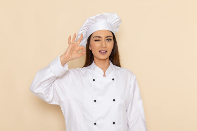 工人身穿白色厨师服的年轻女厨师在浅白的墙上眨眼并显示“好的”标志的正面视图服装年轻好的