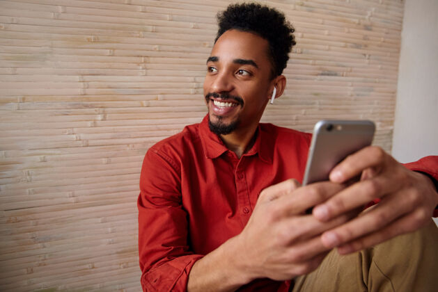 男性年轻快乐的短发胡子黑发男人的肖像 手拿着手机 面带微笑 坐在家里的室内 一边看一边耳机年轻穿着