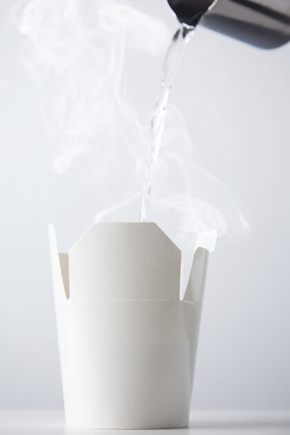 菜肴开水正从一个不锈钢茶壶倒到一个白色纸板拉面盒容器上隔离在白色中国采取外卖