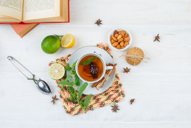 药品一杯肉桂柠檬茶的俯视图 放在方形的柠檬垫上 一碗杏仁 滤茶器和书放在白色的表面上健康香料过滤器