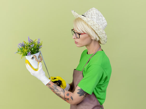 围裙一位年轻的园丁 围裙上留着短发 戴着帽子 手里拿着盆栽植物 拿着卷尺 自信地站在明亮的背景上测量立场头发