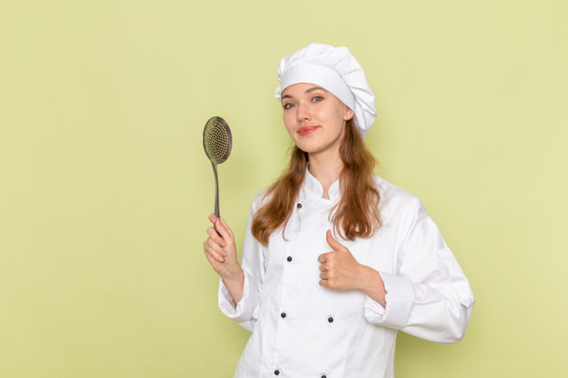 手持身穿白色厨师服的女厨师正拿着大银勺站在绿色的墙上勺子银色烹饪