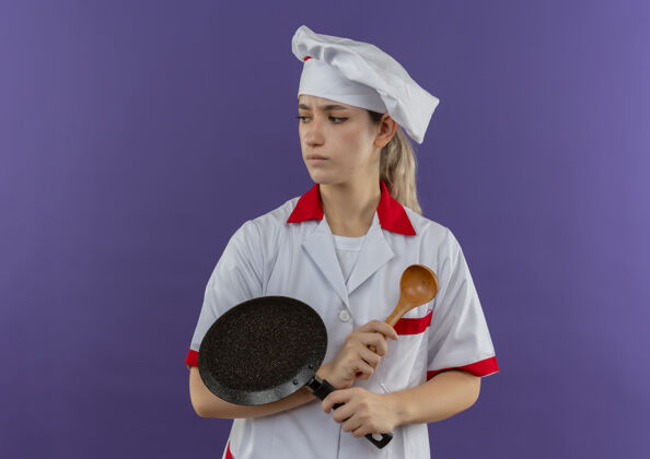 侧年轻漂亮的厨师穿着厨师制服 拿着煎锅和勺子 看着紫色空间里孤立的一面煎厨师勺子