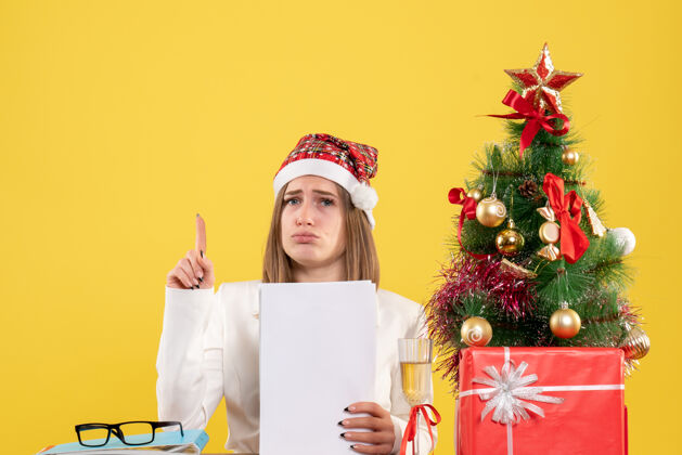 健康正面图：女医生坐在黄色的桌子上 手里拿着圣诞礼物 手里拿着文件前面抱着人