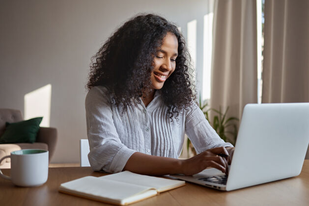 休闲有吸引力的积极的年轻美国黑人女性自由职业者远程工作 在普通笔记本电脑上输入键盘 坐在家里 桌上放着抄写本和马克杯 在网上输入电子信息 面带微笑现代在线快乐