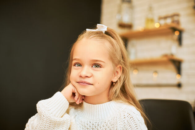 小童年的概念室内形象美丽的长发小女孩坐在厨房里 戴着花环 戴着白丝带和针织毛衣 手放在下巴下 微笑着天真肖像室内