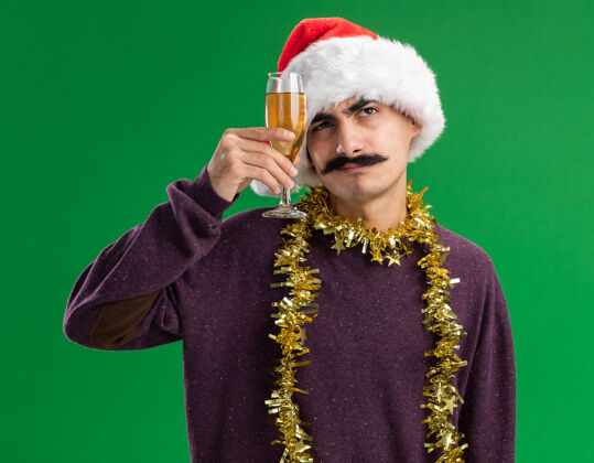 玻璃杯一个留着胡子的年轻人戴着圣诞老人的帽子 脖子上戴着金属丝 手里拿着一杯香槟 困惑地抬头望着香槟脖子金属片