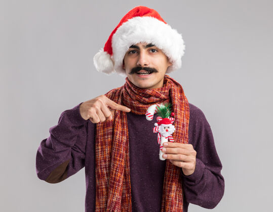圣诞老人快乐的小胡子男人戴着圣诞老人的帽子 脖子上围着暖和的围巾 手里拿着圣诞糖果手杖 用食指指着它微笑着微笑围着快乐