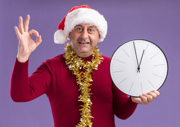 举行中年男子戴着圣诞老人帽子 脖子上戴着金属丝 手里拿着挂钟 微笑着看着相机 紫色背景上立着ok标志墙壁好脖子