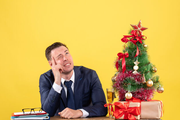坐着前视图思考的人眨着眼睛坐在圣诞树旁边的桌子上 黄色的礼物思考的人办公室桌子