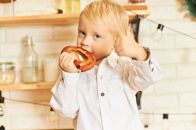男孩食物 烹饪 糕点和面包店的概念英俊的蓝眼睛的小男孩在厨房里享用新鲜出炉的百吉饼 做手势 竖起大拇指标志的肖像手势孩子微笑