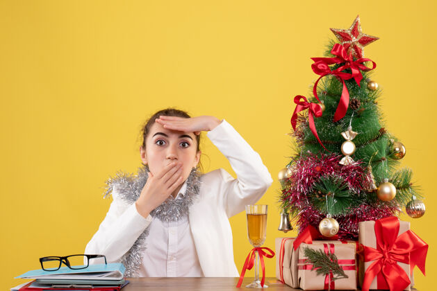 花前视图：女医生坐在黄色背景的桌子后面 带着圣诞树和礼品盒肖像健康快乐
