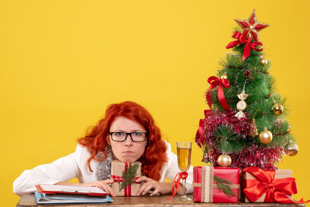 礼物前视图：女医生坐在桌子后面 黄色背景上有圣诞礼物人插花圣诞节