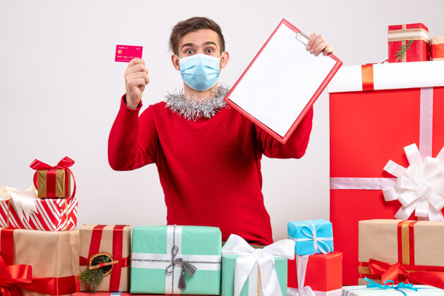 男人前视图年轻人带着面具拿着剪贴板和卡片坐在圣诞礼物周围购物礼物周围