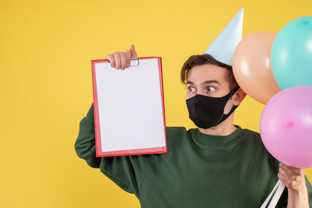 年轻人正面图：戴着派对帽 戴着黑色面具 拿着剪贴板和黄色气球的年轻人男人气球聚会