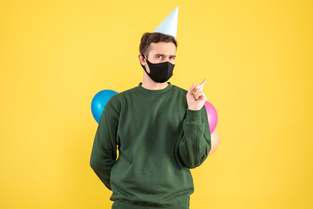 隐藏正面图：戴着派对帽的年轻人和五颜六色的气球把气球藏在背后站在黄色的地板上年轻人人年轻人