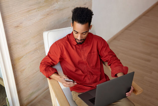 情绪俯视图：年轻的黑发黑皮肤男子身穿红色衬衫 坐在米色内饰的椅子上 拿着笔记本电脑 查看手机上的信息非洲男士坐着