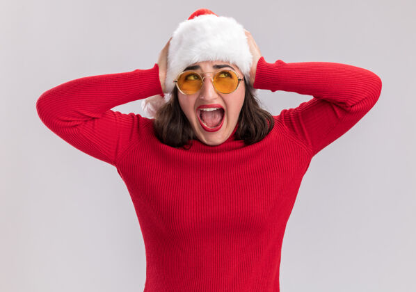 年轻人穿着红毛衣 戴着圣诞帽 戴着眼镜的年轻女孩站在白色的背景下 带着恼怒的表情大声喊叫表情帽子站着